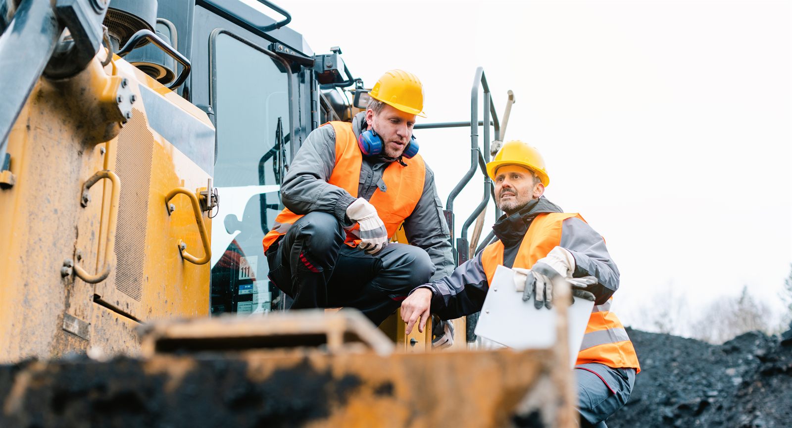 Two site workers in hi-vis and hard hats reviewing a checklist beside heavy earthmoving plant