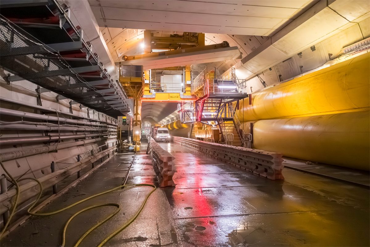 Tunnel lined with segment stacks and a yellow ventilation duct beside a wet haul road